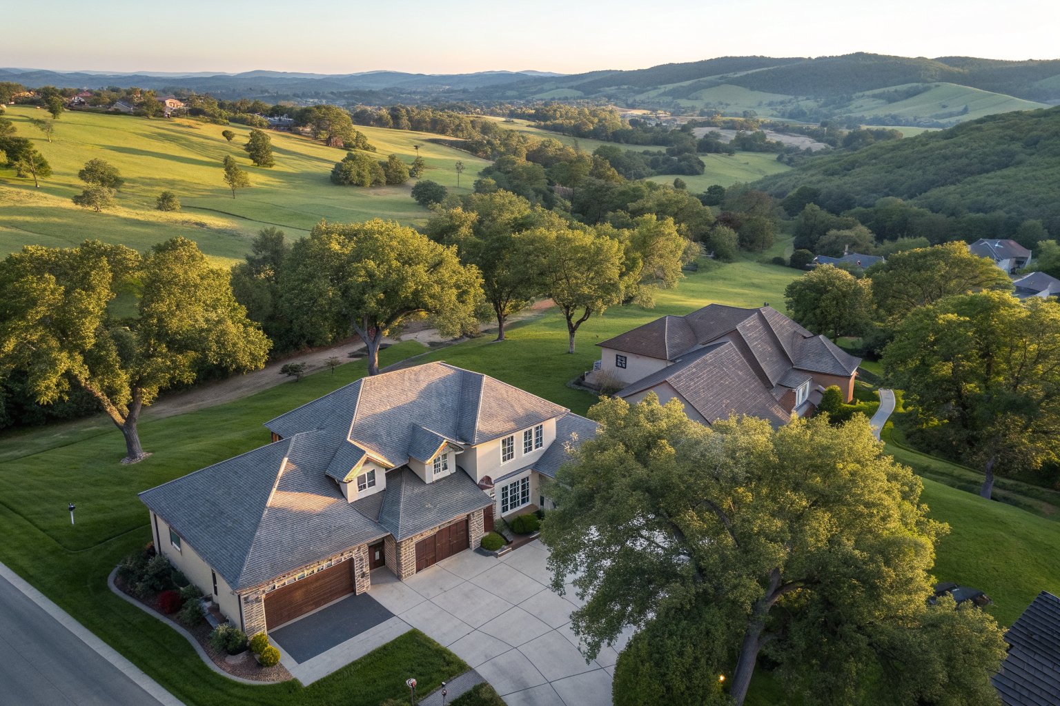 Aerial view of homes with gray and brown shingle roofs among oak trees, green rolling hills beyond