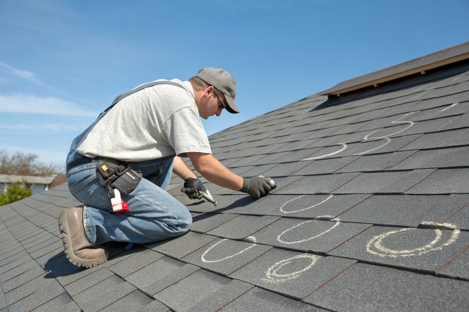 A roofer kneeling on a gray shingle roof checking hail dents circled in chalk